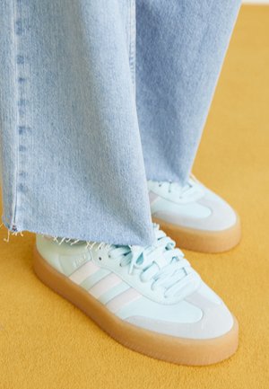 Zapatillas deportivas azul claro con rayas blancas y suelas de goma, usadas con jeans azul claro de dobladillo deshilachado sobre un suelo amarillo.