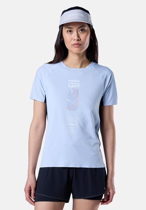 Žena má na sobě světle modré tričko Rossignol s grafikou, černé kraťasy a šedý kšilt Rossignol, stojící před jednobarevným pozadím.