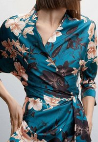 Een teal bloemen wikkeljurk met een diepe V-hals, voorzien van grote perzikkleurige en donkere bloemen, driekwartmouwen en een cinched tailleband.