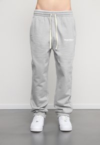 Pantaloni de trening gri, confecționați dintr-un material moale, cu un brâu elastic, șnur, buzunare și un logo "MULTIPLY" în alb.