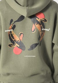 Oliivinvihreä huppari, joka esittelee graafista kuvaa kahdesta koi-kalasta mustana ja oranssina, punaisilla yksityiskohdilla ja valkoisella bränditekstillä alla.