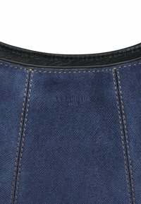 Tissu en denim bleu foncé avec des coutures dorées contrastantes, muni d'un liseré en cuir noir lisse et d'un détail de logo en relief.