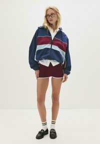Veste coupe-vent colorée en marine, bourgogne et bleu clair, associée à une jupe moulante bourgogne, des chaussettes rayées et des mocassins noirs.
