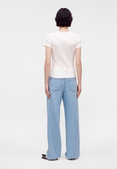Personne aux cheveux courts et foncés portant un t-shirt blanc uni et un jean large bleu clair, debout de dos sur un fond gris clair.