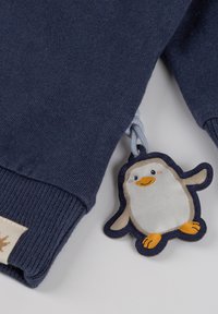 Dunkelblaues Sweatshirt mit gerippten Bündchen und einem penguin-förmigen Stoffetikett, das eine Illustration mit orangefarbigen Akzenten und einer angenähten Schlaufe zeigt.