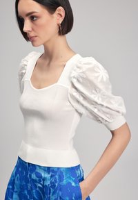 Maglione bianco con corpo testurizzato, scollatura quadrata e maniche trasparenti a palloncino con ricami floreali. Abbinato a pantaloni blu con stampa.