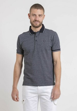 Homme aux cheveux courts et barbe portant un polo bleu marine à motifs avec un col noir et un pantalon blanc sur un fond blanc uni.