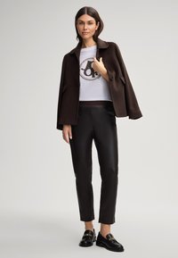 Bruine wollen jas over een witte cropped t-shirt met logo, gecombineerd met zwarte leren broek en zwarte loafers met gouden hardware accenten.