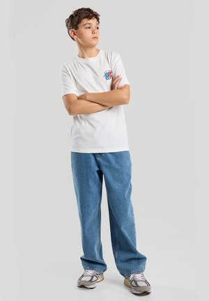 White cotton t-shirt with blue and red graphic on the chest, paired with blue denim jeans and gray sneakers featuring white accents.