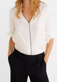 Blouse blanche à col en V avec manches retroussées, ornée d'un passepoil noir le long de la patte de boutonnage. Portée avec un pantalon noir, créant un contraste.