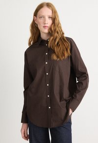 Brown button-up shirt in cotton, featuring a pointed collar, long sleeves, and a chest pocket. Notable white buttons and an oversized fit.