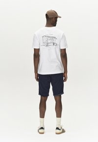 T-shirt bianco con un'illustrazione in outline nera sul retro, abbinato a pantaloni corti blu scuro e calzini chiari. Uomo con un cappello marrone.
