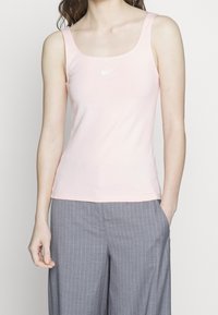 Pink cotton tank top with a scoop neckline, thin straps, and a small white Nike logo on the chest. Paired with grey striped trousers.