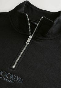 Black pullover with a quarter-zip collar. Features a silver zipper, soft texture, and embroidered "BROOKLYN" design in blue.