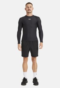Zwarte lange mouwen sportshirt met ronde hals, gecombineerd met zwarte shorts. Bevat een logo op de borst, gedragen met witte sokken en zwarte schoenen.