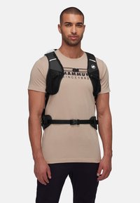 Zwarte sportharnas met meshsecties, verstelbare banden en een centrale gesp. Draagt over een lichtbeige T-shirt met een logo en tekst.