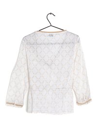 Blusa blanca semitranslúcida de manga larga con patrón de diamantes y ribetes beige en los puños y el escote, colgando de una percha negra sobre un fondo blanco.