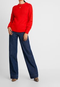 Pull en tricot rouge avec un motif texturé complexe sur les épaules, associé à un jean noir à taille haute et à pattes d'éléphant, et à des talons à motif.