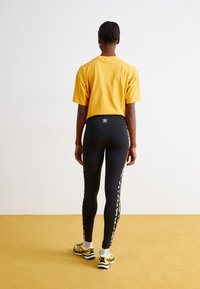 Fekete leggings leopárd mintás oldalrésszel, sárga rövid ujjú felsővel és arany tornacipővel párosítva, lazán illeszkedő stílusban.