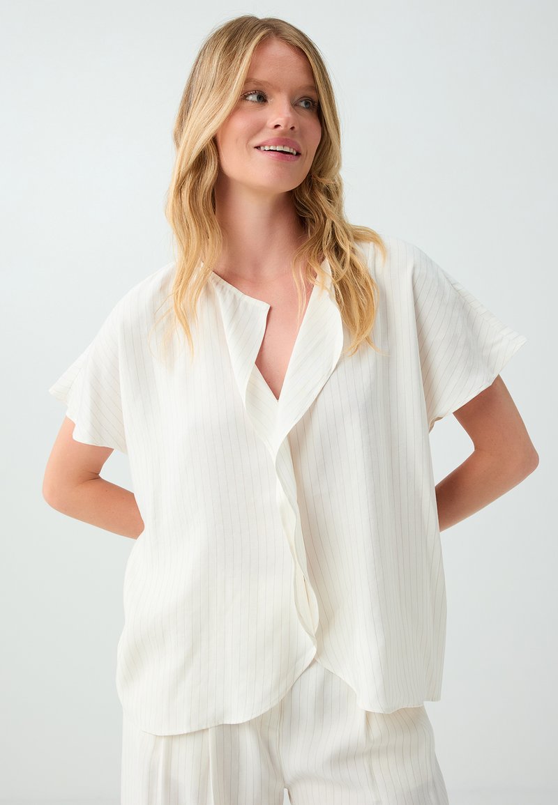 Femme blonde portant une chemise blanche ample à manches courtes et un pantalon assorti, souriante et regardant sur le côté sur un fond uni.