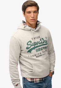 Felpa con cappuccio grigio chiaro con coulisse, con logo e scritta "SuperDry" verde. Indossata sopra una camicia a quadri al colletto.