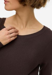 Tumši brūns ribots džemperis ar plašu izgriezumu un garām piedurknēm, tuvplāns ar roku, kas atpūšas pie apkakles. Gluda tekstūra.