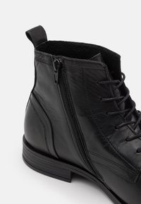 Botín de cuero negro hasta el tobillo con cierre de cremallera, diseño de cordones, textura suave y suela resistente con un ligero tacón.