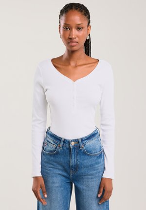 Junge Frau mit geflochtenen Haaren, die ein weißes langärmliges Oberteil und hoch taillierte blaue Jeans trägt, vor einem schlichten Hintergrund steht.