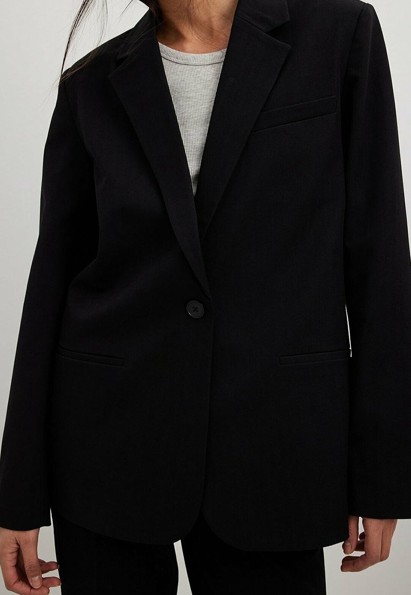Personne portant un blazer noir à un seul bouton sur une chemise côtelée gris clair, à manches longues et poches avant, raccourcie au niveau du cou et des hanches.