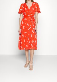 Robe portefeuille rouge à fleurs avec des manches courtes volantées, un décolleté en V et une taille nouée. Le tissu est léger et semi-transparent.