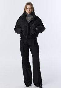 Doudoune noire, coupe courte avec col montant, dotée d'une ceinture côtelée. Le pantalon est large et noir.