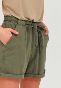 Olive green shorts made of textured fabric, featuring an elastic waistband with a drawstring and side pockets. Rolled cuffs at the hem.