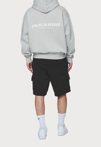 Felpa grigia con testo "PEGADOR" bianco, pantaloni cargo neri, sneakers bianche e calzini corti, visti da dietro su uno sfondo neutro.