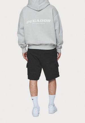 Szary sweatshirt z białym napisem "PEGADOR", czarne szorty cargo, białe trampki i skarpetki do kostek, widok od tyłu na jednolitym tle.