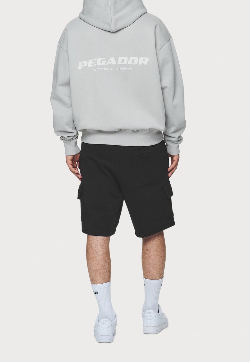 Felpa grigia con testo "PEGADOR" bianco, pantaloni cargo neri, sneakers bianche e calzini corti, visti da dietro su uno sfondo neutro.