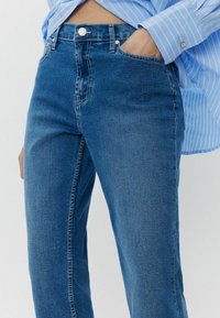 Siniset farkut korkealla vyötäröllä, joissa on etutasku, vyölenkit sekä klassinen nappi- ja vetoketjukiinnitys.