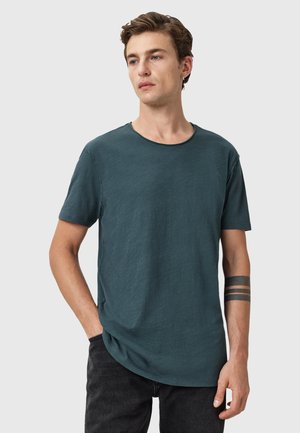 Camiseta de manga corta en verde azulado oscuro con cuello redondo, con un corte ligeramente relajado y tela texturizada. Detalle de costura notable en el escote.