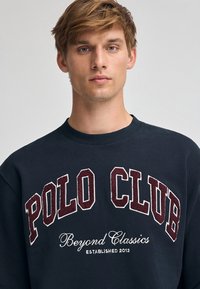 Mörkblå sweatshirt med "POLO CLUB" i borgundy texturerad lettering och "Beyond Classics" i vit kursiv, design med rund hals.