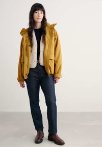 Geel parka met capuchon over een beige fleecvest, gecombineerd met donkerblauwe jeans en bruine enkellaarsjes. Soepele textuur, casual pasvorm.