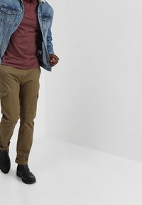 Muž v hnědých cargo kalhotách, černých botách, šarlatové košili a modré džínové bundě kráčející proti jednolité šedé pozadí.