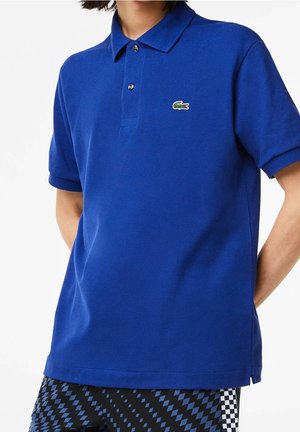 Osoba ubrana w niebieską koszulkę polo Lacoste w połączeniu z czarno-niebieskimi spodniami w kratę.