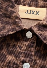 Chemise en velours côtelé marron avec une texture douce, un col et un bouton-pression argenté, portant une étiquette affichant la marque "JJXX" en noir.