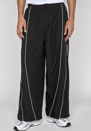 Schwarze, weit geschnittene Trainingshose mit weißen Paspeln, getragen mit weißen und grauen Sneakers, an einer Person von der Taille bis zu den Füßen gezeigt.