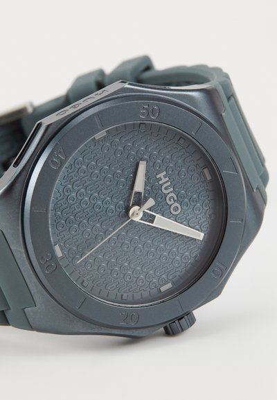Montre Hugo avec un cadran texturé bleu foncé, aiguilles et index argentés, bracelet en silicone et un boîtier rond avec une lunette surélevée.