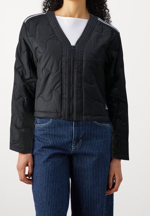 Personne portant une veste matelassée noire à fermeture éclair sur un haut blanc et un jean bleu à fines rayures sur un fond blanc uni.