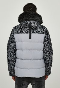 Giacca imbottita tipo puffer con cappuccio e maniche a motivo geometrico nero, corpo grigio e rifinitura in pelliccia attorno al collo. Presenta un dettaglio con fibbia sul retro.