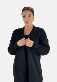 Svart kardigan med ribbad textur, öppen fram, och överdrivna, texturerade ärmar med ett kabelstickat mönster.