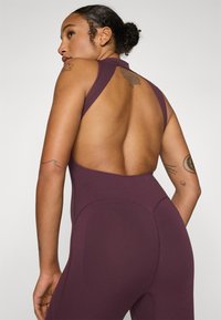 Burgundy athletic jumpsuit with a backless design, featuring wide straps and a textured finish. Accentuates body shape with curved seams.