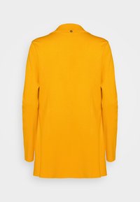 Cappotto giallo su misura con una silhouette elegante, caratterizzato da un colletto alto e un unico bottone sul retro, realizzato in un tessuto liscio e texture.