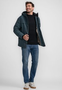 Mørk teal polstret jakke med hætte, båret over en sort ribbede sweater, parret med lyseblå jeans og grå sneakers med brune såler.